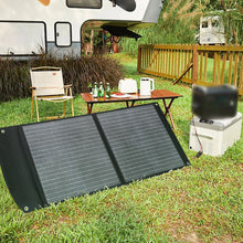 Load image into Gallery viewer, SolarOutside 20W portable monocrystalline solar panel charger unfolded in a grassy RV camping site. A large black solar blanket is set up on the lawn, ready to power portable energy storage and a mini fridge near a parked camper trailer and outdoor seating area.