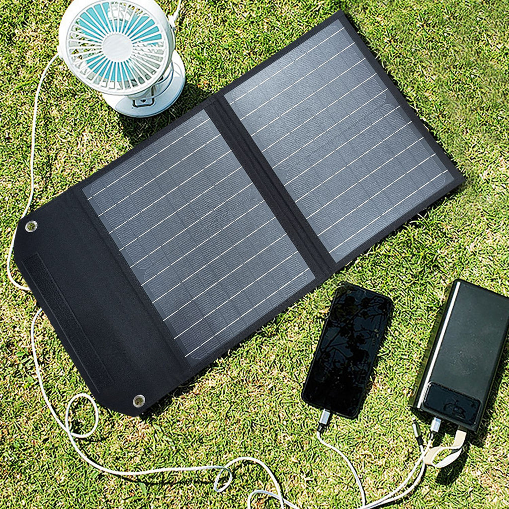 SolarOutside 20W portable solar panel charger laid flat on a grassy lawn. The monocrystalline solar bag is actively charging a smartphone, a high-capacity power bank, and a small rechargeable fan simultaneously using its built-in USB output.