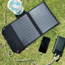 Load image into Gallery viewer, SolarOutside 20W portable solar panel charger laid flat on a grassy lawn. The monocrystalline solar bag is actively charging a smartphone, a high-capacity power bank, and a small rechargeable fan simultaneously using its built-in USB output.